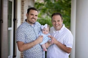 Two dads smiling outside with their baby daughter