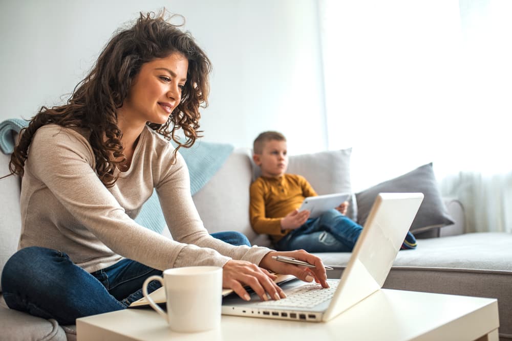 Woman working on a laptop from a couch organizing adoption records in Texas with a young male child in the background holding a digital tablet