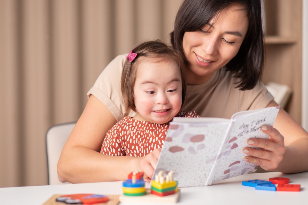cute child with down syndrome girl with mom and book at home See less
