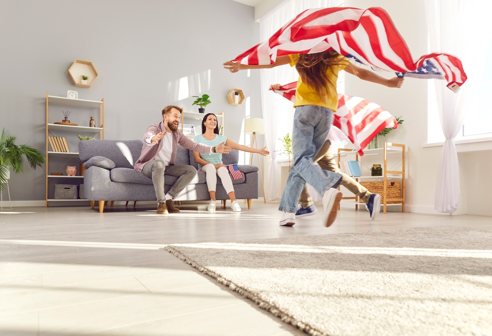 Happy funny children boy and girl having fun and running with flags of united states in living room at home celebrating Independence Day with their parents. Family patriotism and US holiday concept