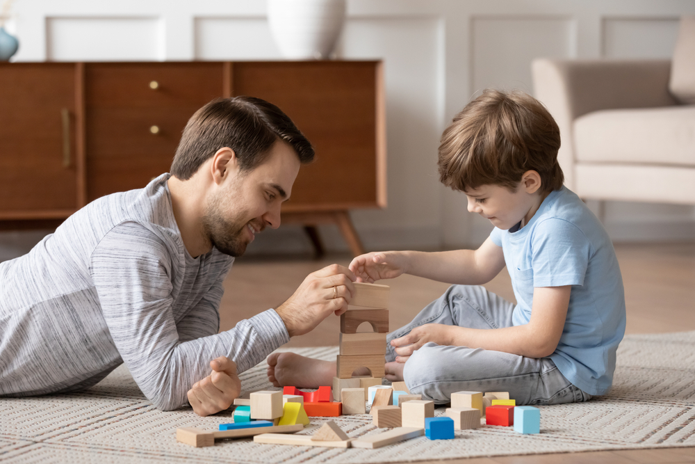 Loving young father lying on floor in living room playing building bricks with little preschoolers son, caring dad relax with small boy child engaged in funny learning activity at home together