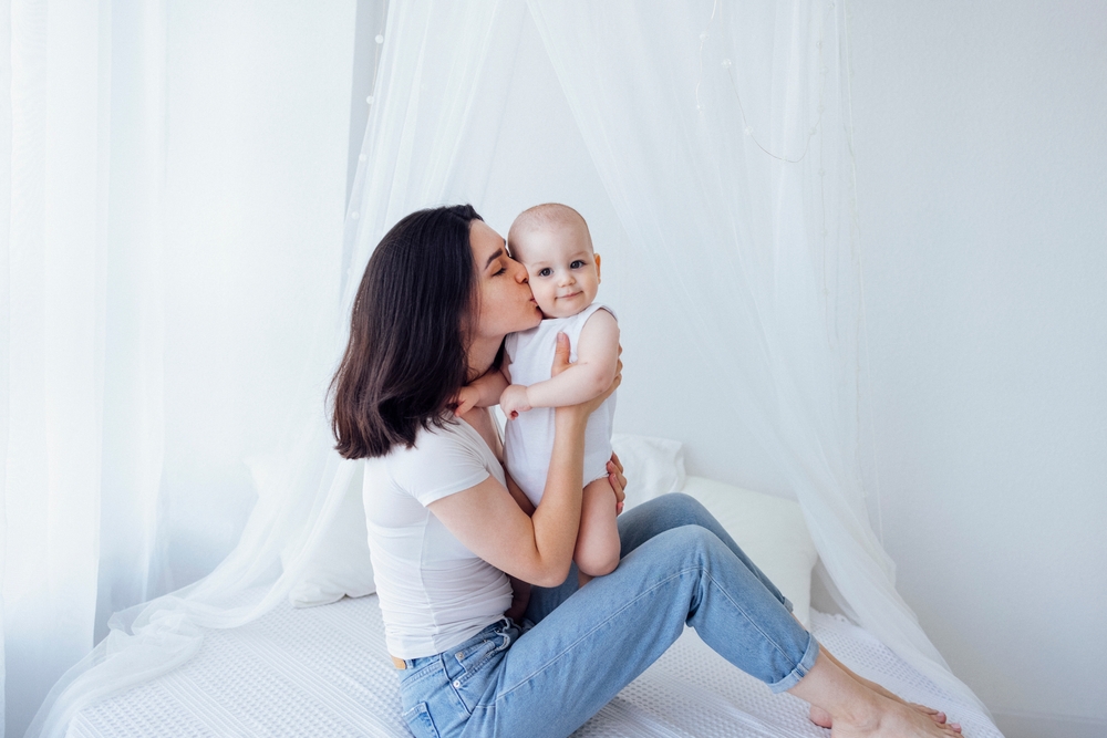 Young beautiful mother kisses her cute baby on cheek. White brunette touches her smiling child with her face. Pretty infant in a white bodysuit with his mom on a light background. Motherhood concept.